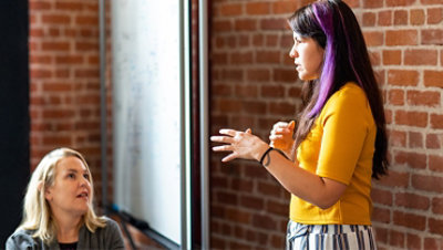 A female developer is speaking near a white board during team stand up meeting.