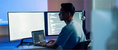 V slabo osvetlenej miestnosti sedí za stolom osoba, ktorá pracuje na troch počítačových obrazovkách zobrazujúcich kód.