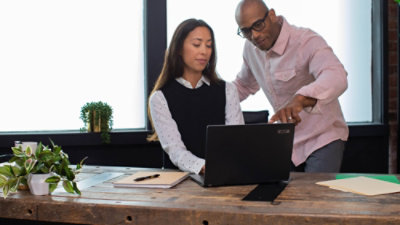 Deux personnes collaborent sur un bureau en bois et examinent le contenu d’un ordinateur portable dans un espace de travail lumineux avec des plantes et des fournitures de bureau à proximité.