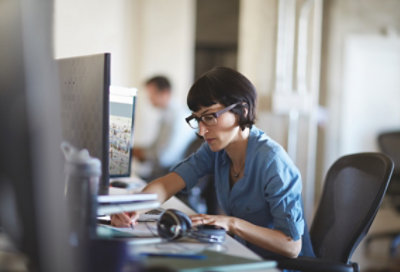 A person writing something by hand while seated at a desk with two large desktop monitors.