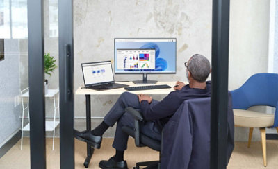 Een persoon die in een glazen kantoor zit en een laptop gebruikt die is verbonden met een desktopmonitor