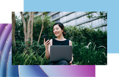 En smilende kvinne med grått krøllete hår som ser på smarttelefonen sin og holder briller, står på et fortau i byen.