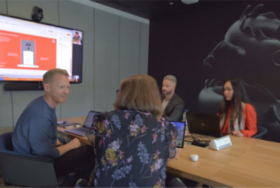 Fire personer i et mødelokale er engageret i et videoopkald og en præsentation med åbne bærbare computere og en skærm, der viser slides.