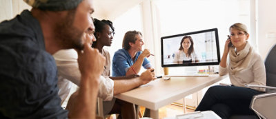 Cztery osoby siedzą przy stole i uważnie wpatrują się w ekran komputera wyświetlający rozmowę wideo z inną osobą.