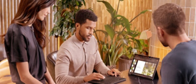  man sitting at a desk with a laptop and two other people standing.