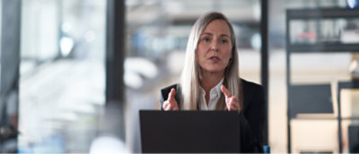 Una persona con capelli lunghi e un'espressione seria seduta a una scrivania con un portatile, che gesticola con entrambe le mani, in un ufficio moderno.