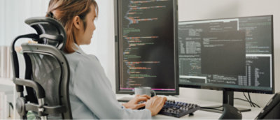 Eine Person befindet sich an einer Doppelmonitoreinrichtung in einem Bürostuhl und gibt Code auf ihrer Tastatur ein.
