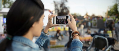 Une personne prend une photo avec un smartphone pendant un événement en plein air, avec d’autres personnes et des décorations colorées visibles en arrière-plan.