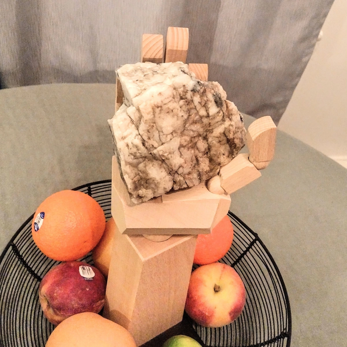 A life-sized wooden hand on a stand holding a baseball-sized white, semi-translucent rock with black veins. The hand rests in a fruit bowl on a table. There are peaches, clementines, asian pears and a lime in the bowl.