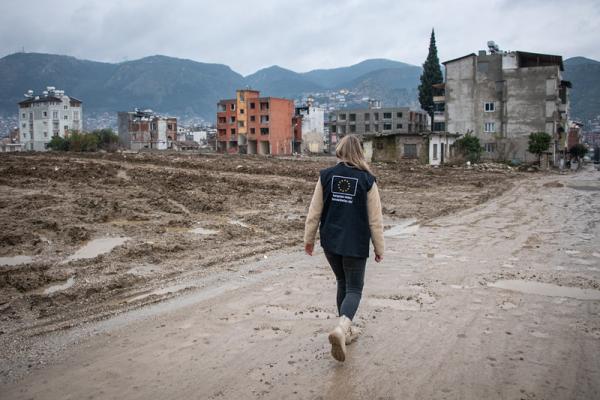An aid worker wearing a dark blue vest with the European Union humanitarian aid emblem walks along a muddy, debris-strewn street through a devastated urban area of partly ruined buildings, with misty mountains rising in the background.