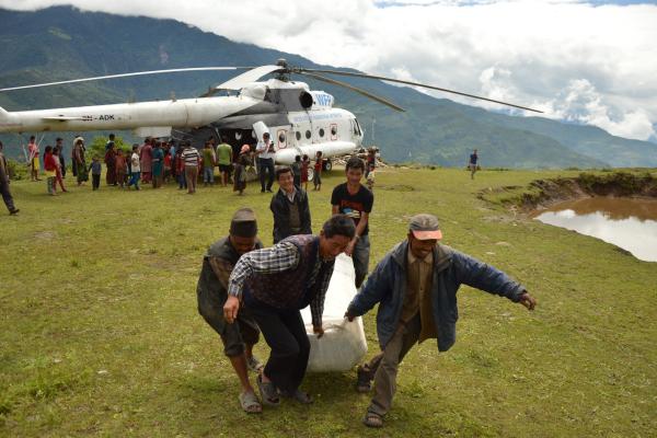 In a remote, grassy landing area in the mountains, a large white helicopter marked with the World Food Programme logo is parked while a crowd of villagers queues beside it. In the foreground, four men with blurred faces struggle together to carry a heavy, wrapped sack away from the helicopter, suggesting the unloading and distribution of humanitarian relief supplies in a rural community.