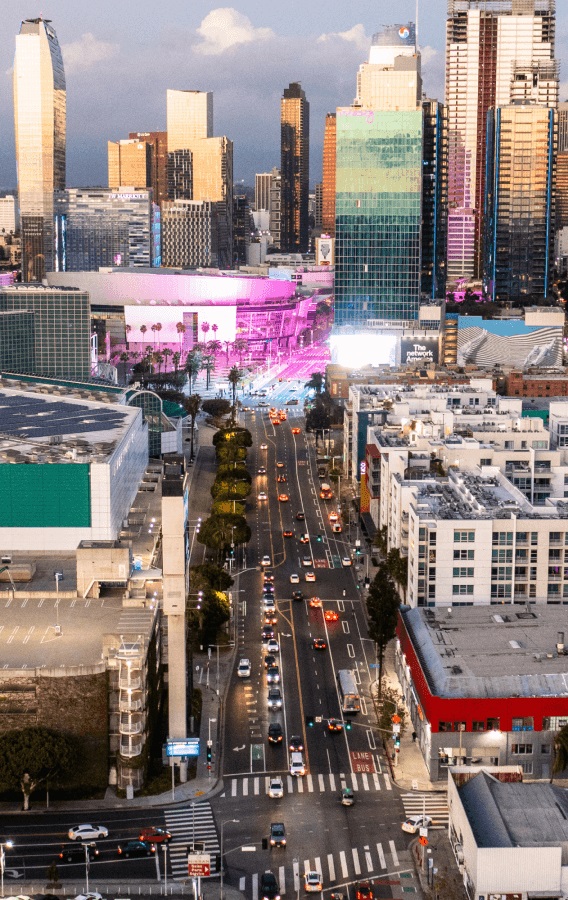 Aerial view of downtown Los Angeles at dusk, featuring skyscrapers, a brightly lit arena with pink lights, and a busy street with cars.