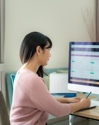 A person wearing a pink sweater is seated at a desk, writing in a notebook while looking at a computer screen displaying a website interface.