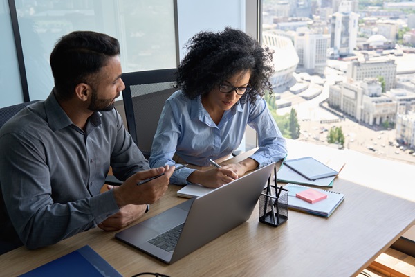 Two people working together at a desk with a laptop, notebooks, and a cityscape visible through a large window.