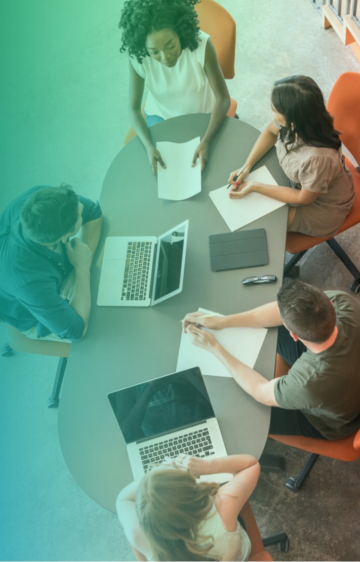 A group of people sitting around a table with laptops, papers, and a tablet, engaged in a discussion.