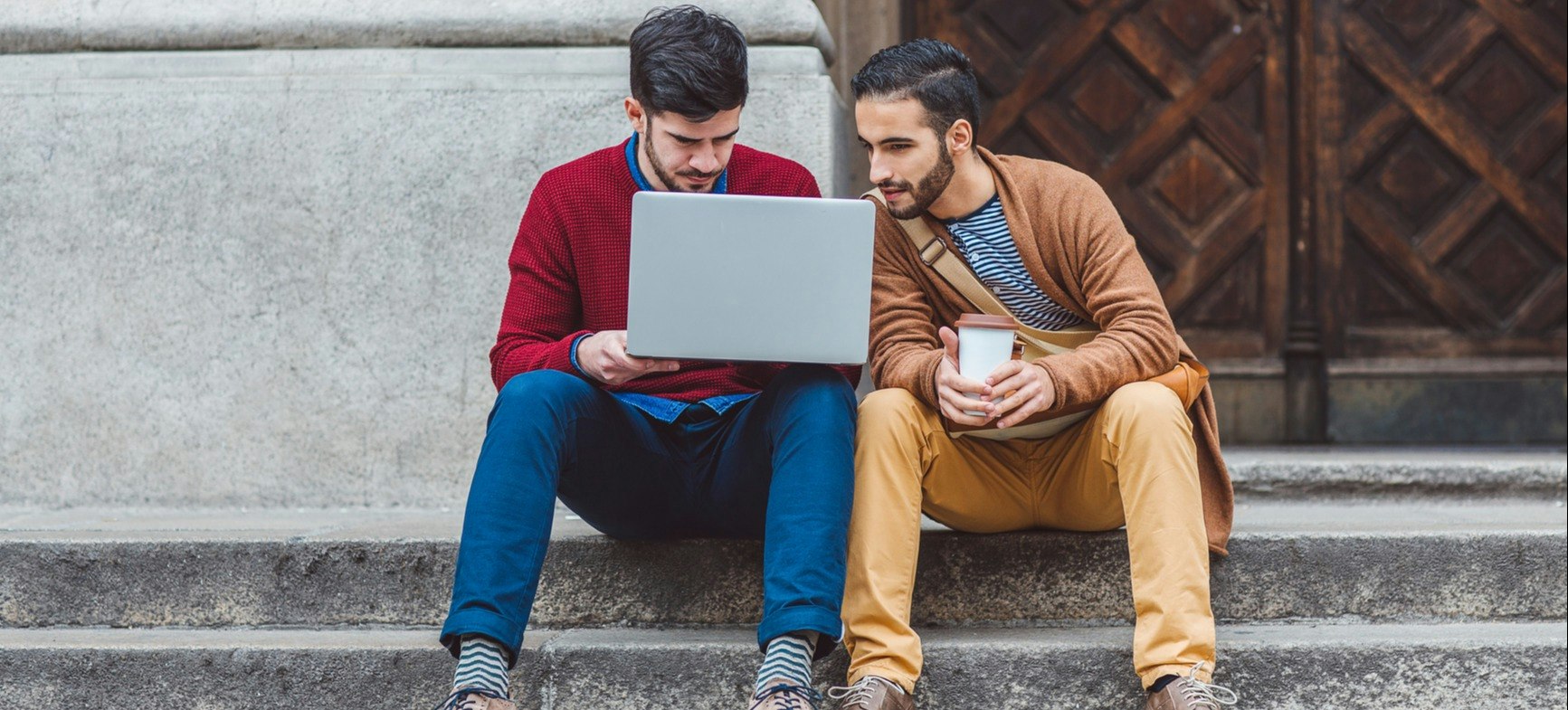 [featured image] Two young men wearing colored sweaters sit on the steps outside of a building collaborating on a laptop. 