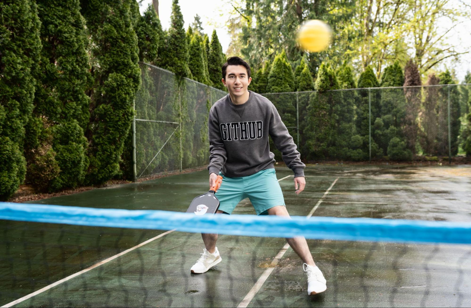 A smiling man wielding a pickleball paddle is wearing a grey sweatshirt with