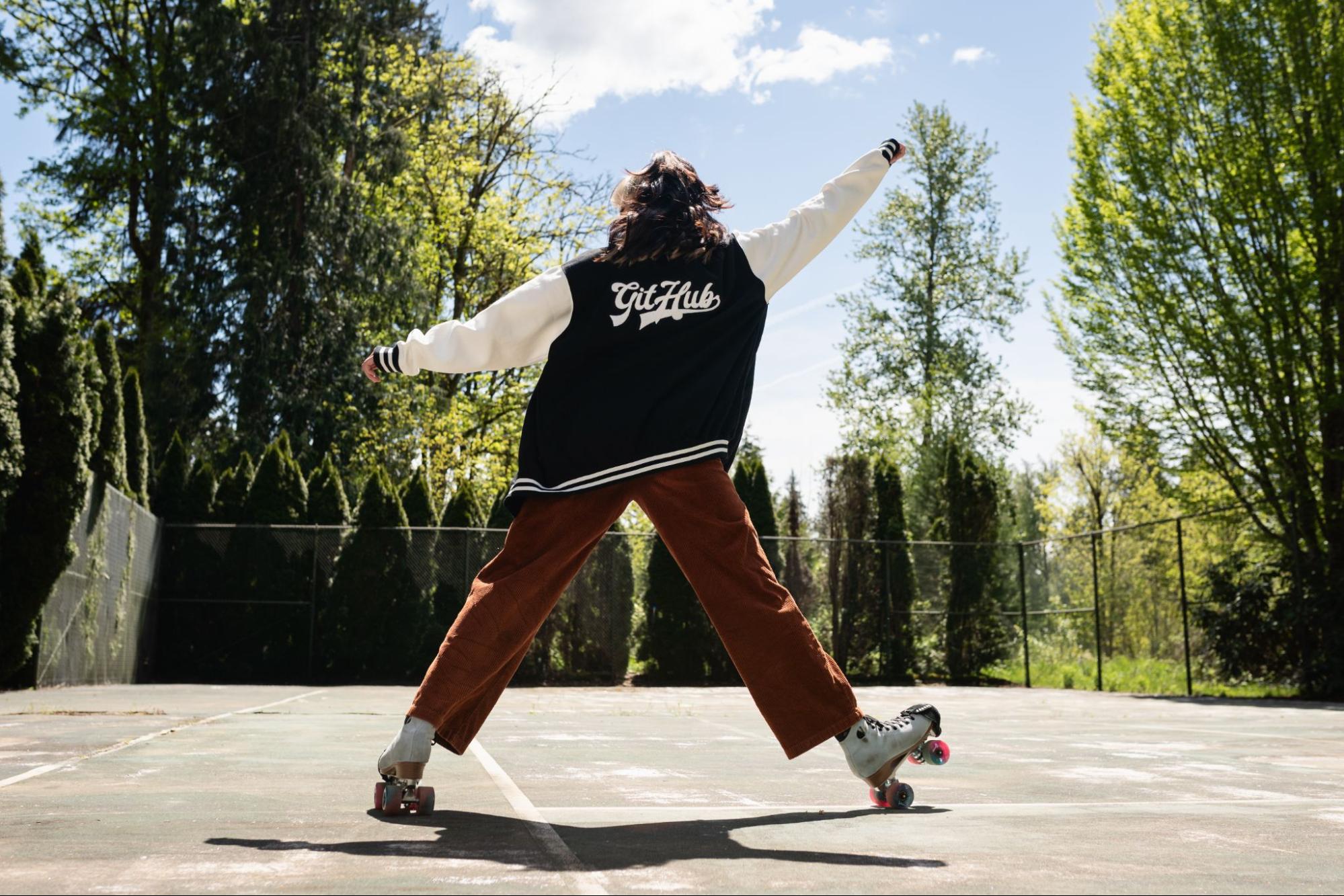 A woman wearing roller skates has her back to the camera and her arms in the air in a triumphant pose. She is wearing a varsity letter style jacket that says