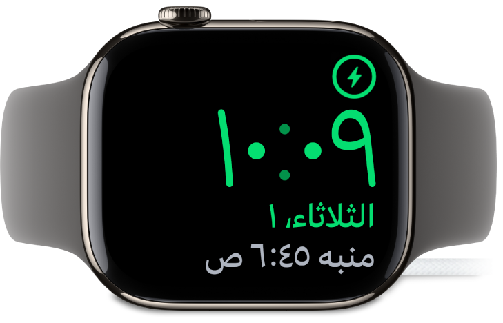 Apple Watch موضوعة على جانبها ومتصلة بالشاحن، وتعرض الشاشة رمز الشحن في الزاوية العلوية اليمنى، والوقت الحالي أسفل ذلك، ووقت المنبه التالي.