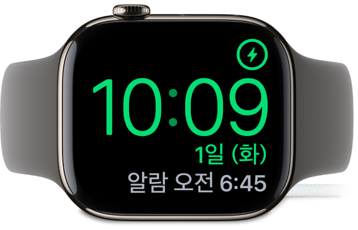 Apple Watch가 옆으로 누워 있고 충전기에 연결되어 있음. 화면의 오른쪽 상단 모서리에는 충전 기호가 있고 그 아래에는 현재 시간과 다음 알람 시간이 있음.