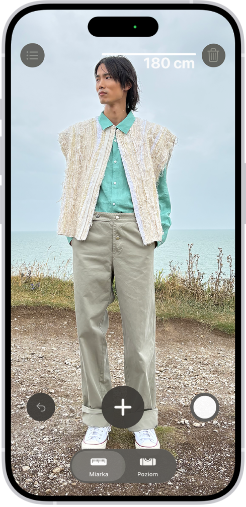 Pomiar wzrostu osoby w aplikacji Miarka. Nad głową mierzonej osoby wyświetlany jest zmierzony wzrost. W prawym dolnym rogu widoczny jest przycisk migawki, który pozwala zrobić zdjęcie pomiaru. U góry ekranu widoczny jest zielony wskaźnik użycia aparatu.