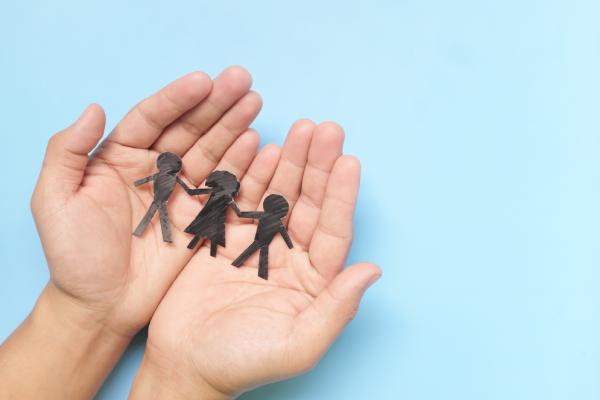 The visual shows two open and juxtaposed hands that hold inside a cut-out of paper representing children, as they would be drawn by a child. Against a light blue background, the image suggests the protection of children.
