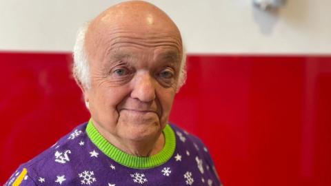 An older man looking into the camera wearing a purple jumper with a green trim