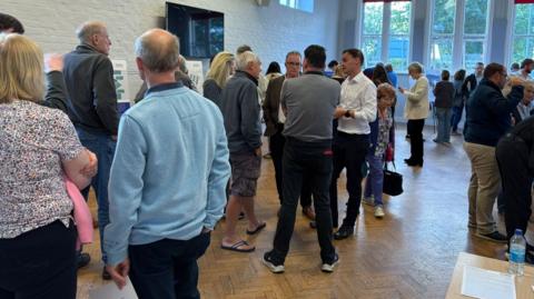 A village hall with painted white brick interior and wooden floor, with a gathering of people all chatting.