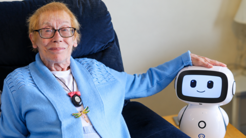 Vera, a 74-year-old wearing glasses and a blue cardigan with a dragonfly brooch, rests her hand on Robert the robot's head