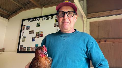A man in a red cap, wearing black framed glasses, is holding a chicken under one arm. There is a board behind him that says feathered friends