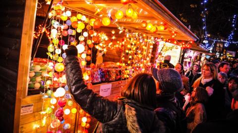 A crowd of people in thick winter clothes at night time are looking towards a well lit wooden cabin, which has various items hanging from it seemingly for sale.