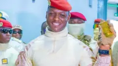 Ibrahim Traoré in military uniform and a red beret smiles while holding up a gold bar in his gloved hand. Several uniformed officers stand behind him