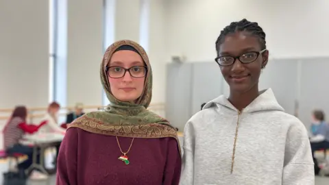 Two teenage girls look directly at the camera. One is wearing a grey hoodie and the other is wearing purple jumper and a headscarf. 