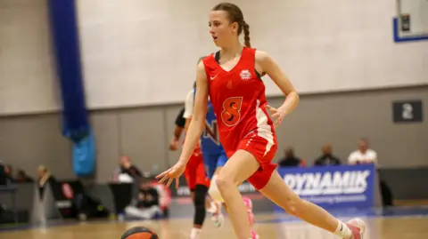 Basketball England Jess, 15, playing basketball in a high-level competitive tournament. She is wearing a red jersey and shorts and is dribbling the ball.