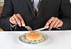 A suited person cuts a burger placed on a dollar bill with a knife and fork.