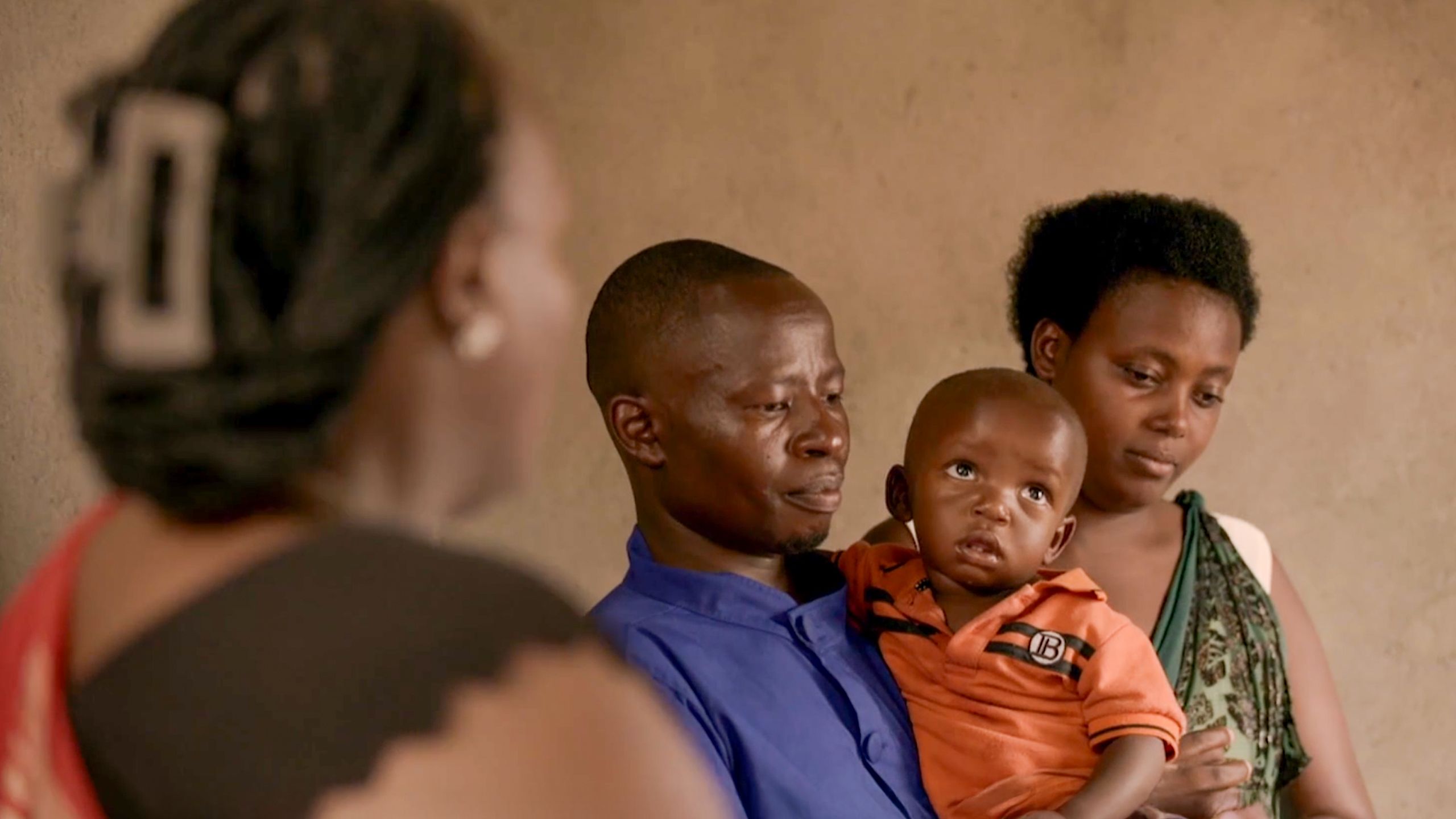 A man holding a baby and a woman nearby, a person in the foreground is blurred.