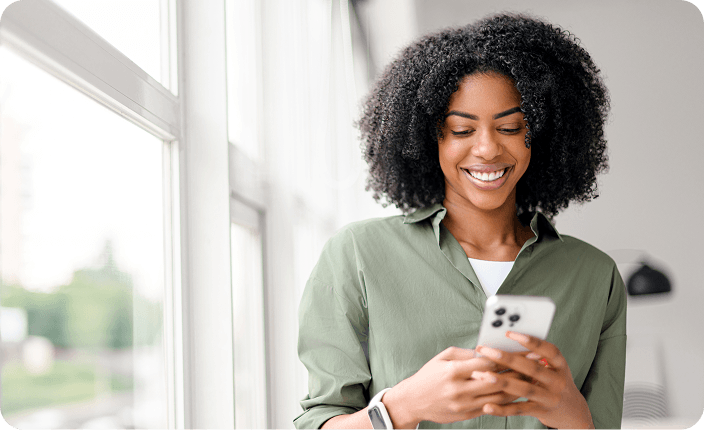 Une femme souriante qui regarde son téléphone intelligent près d'une fenêtre lumineuse.