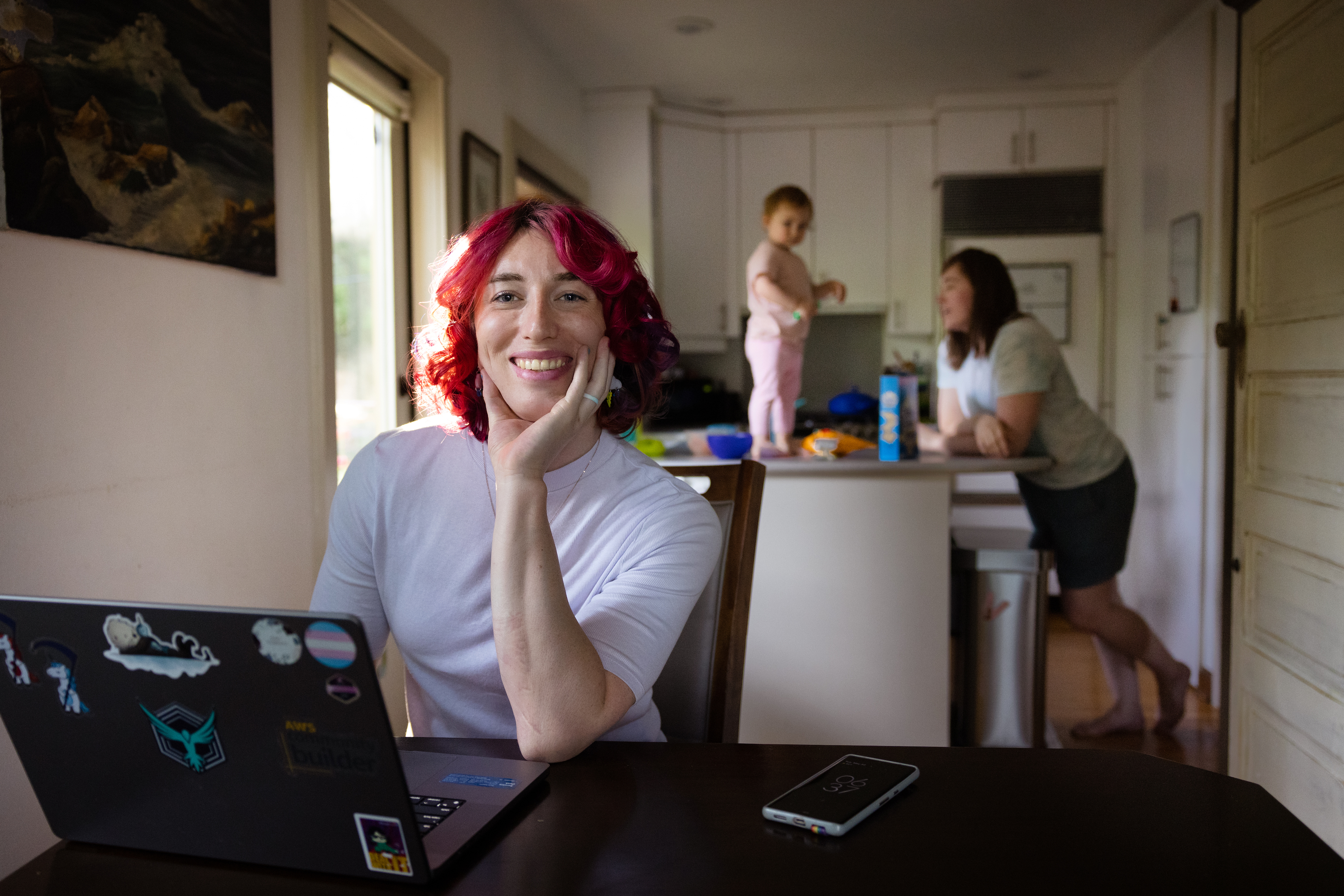 Kyler smiles as she sits at her desk in the kitchen, with her daughter and partner in the background.