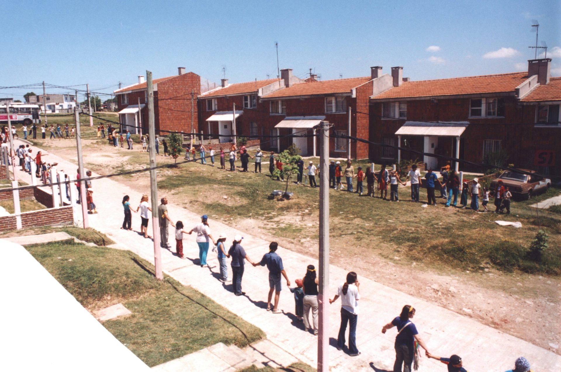 Children and adults hold hands along a sidewalk in front of low-rise homes.