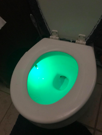 A different reviewer image showing their toilet seat shining bright green in a room with the lights turned on