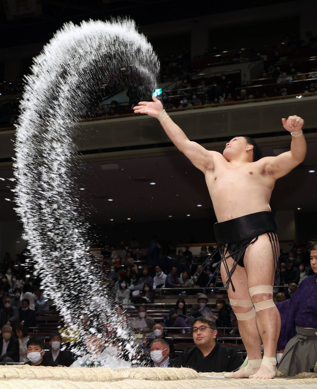 男性力士が土俵上で塩を撒いて儀式を行っている様子。観客が背景に見える。