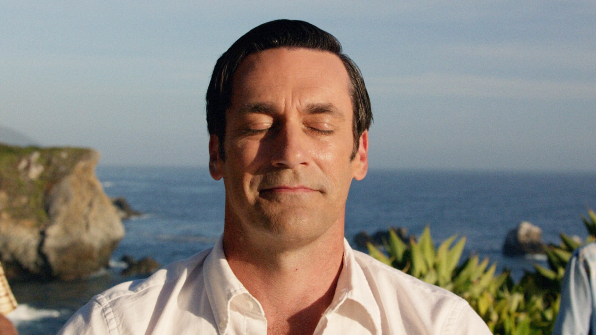 Person with eyes closed, outdoors near ocean, wearing a collared shirt; appears serene and contemplative