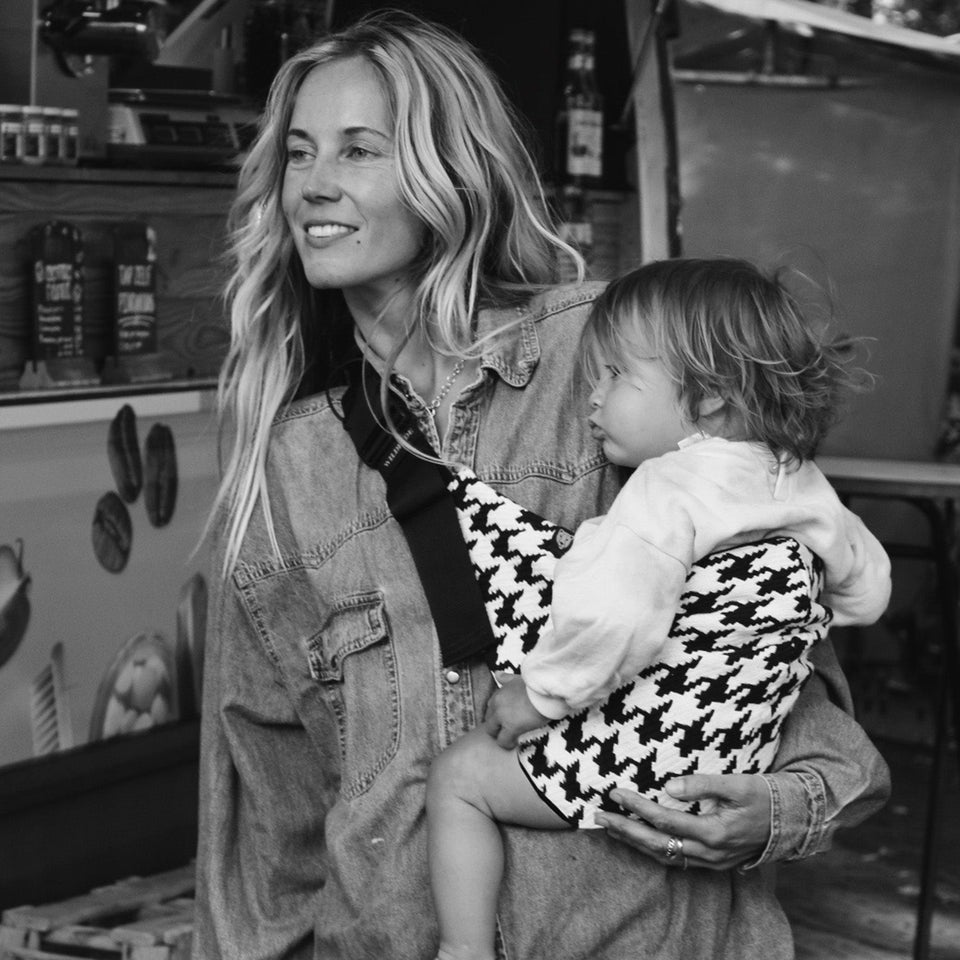 Person in denim jacket holding a small child wearing a patterned outfit. Background shows outdoor market stalls