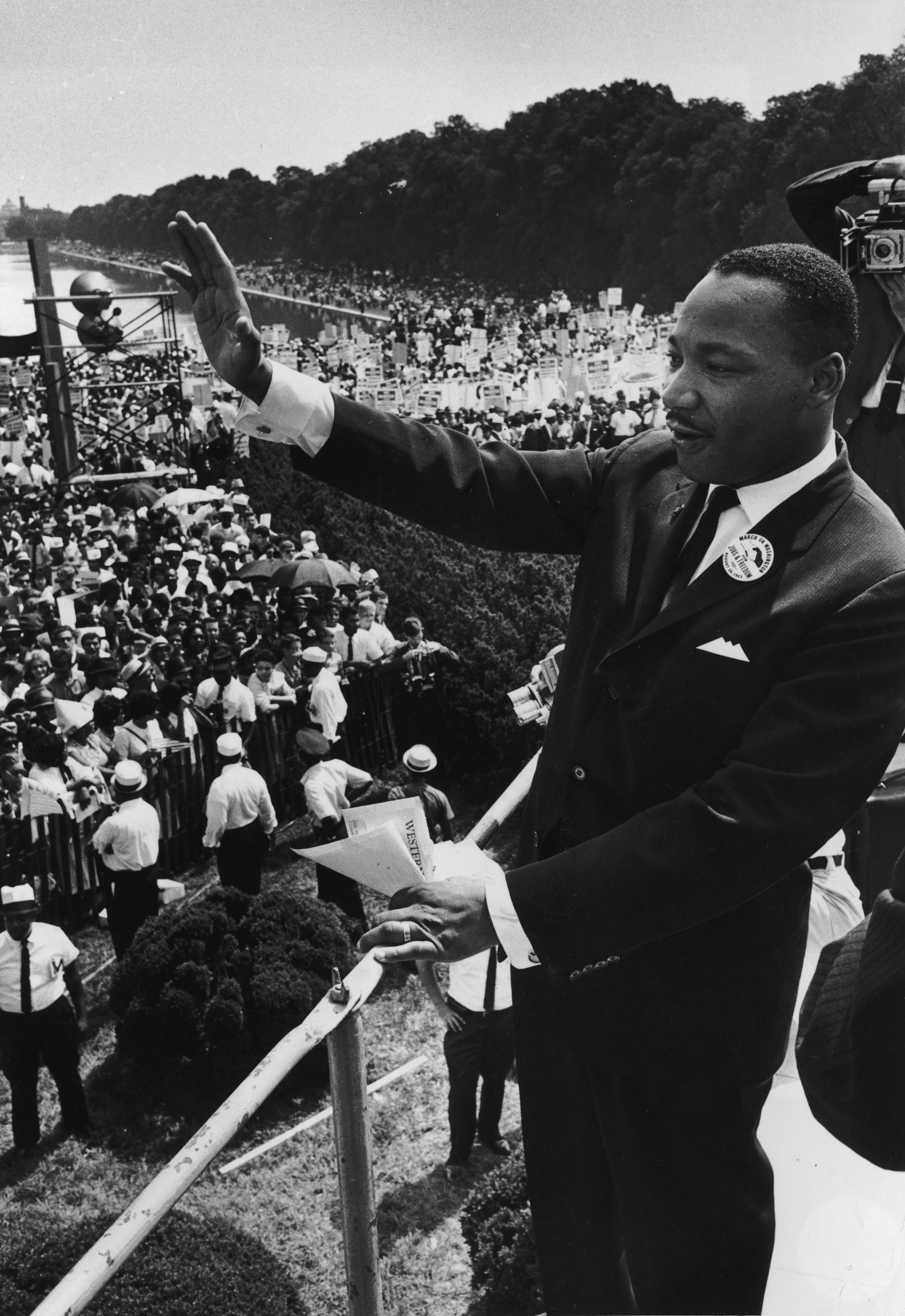Martin Luther King Jr. waves to a large crowd at the 1963 March on Washington, standing on a platform holding papers