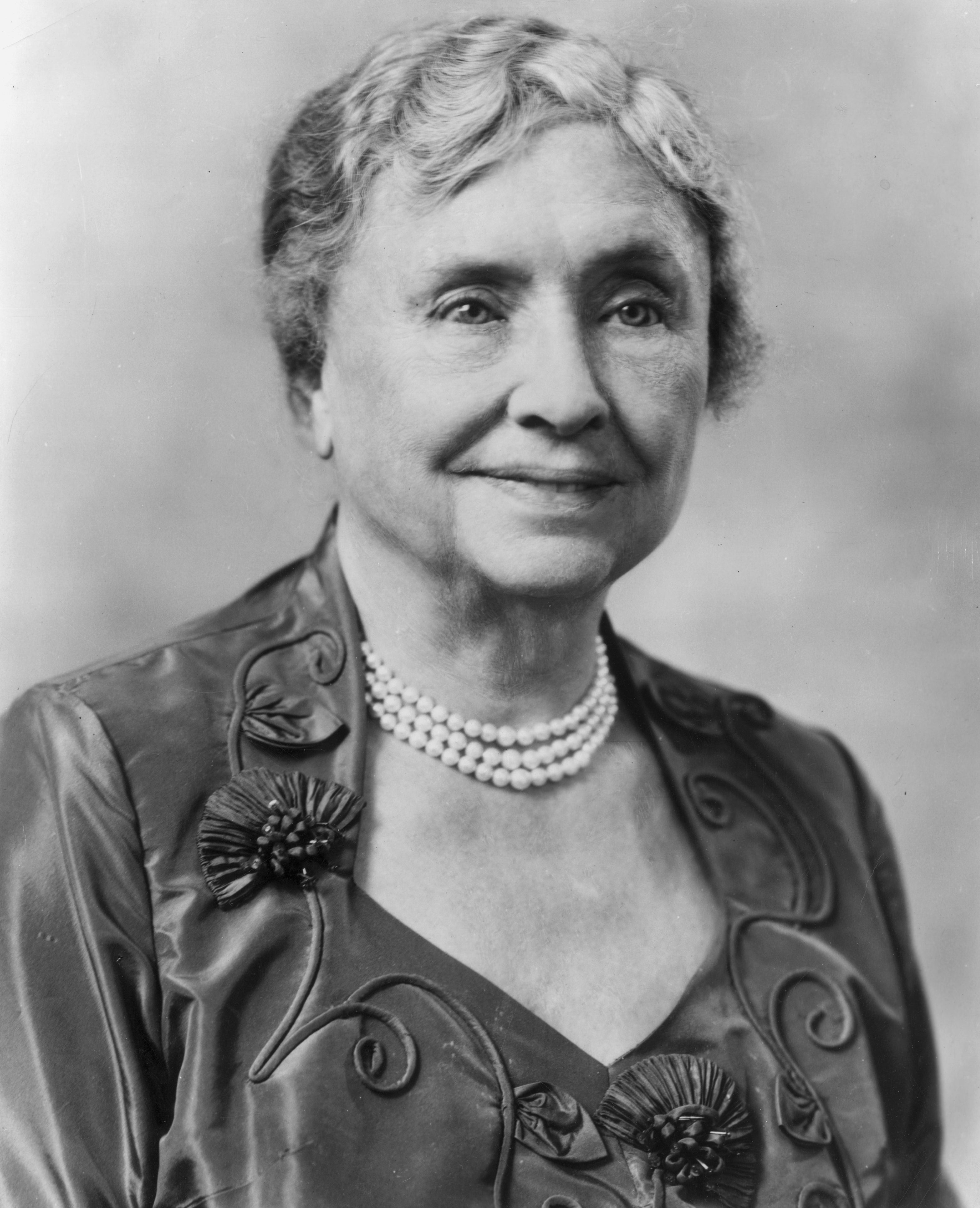 An older woman with short hair, wearing a dress with floral details and a pearl necklace, smiles softly at the camera