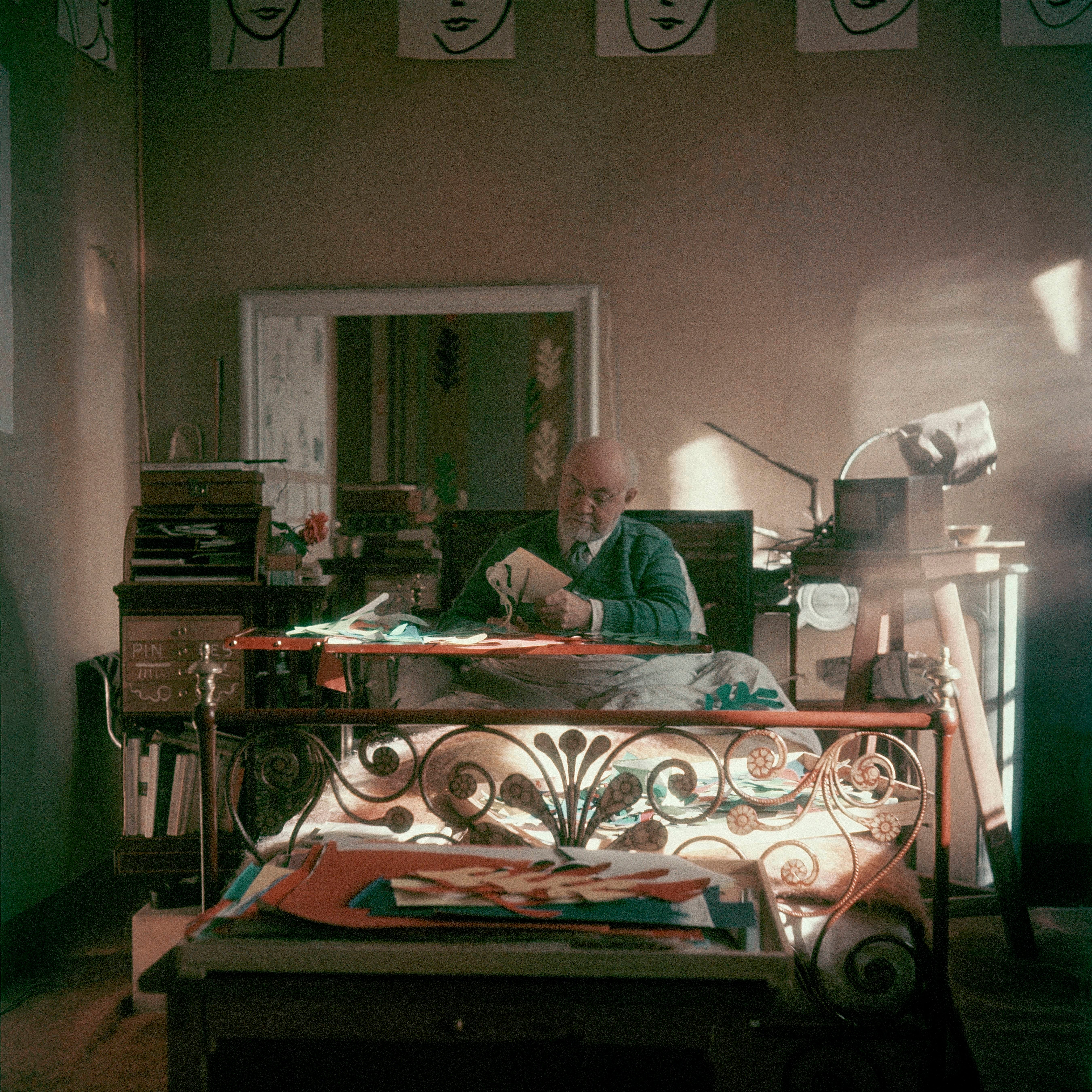 An elderly person works with paper cutouts at a desk in an artistic studio filled with books and art supplies