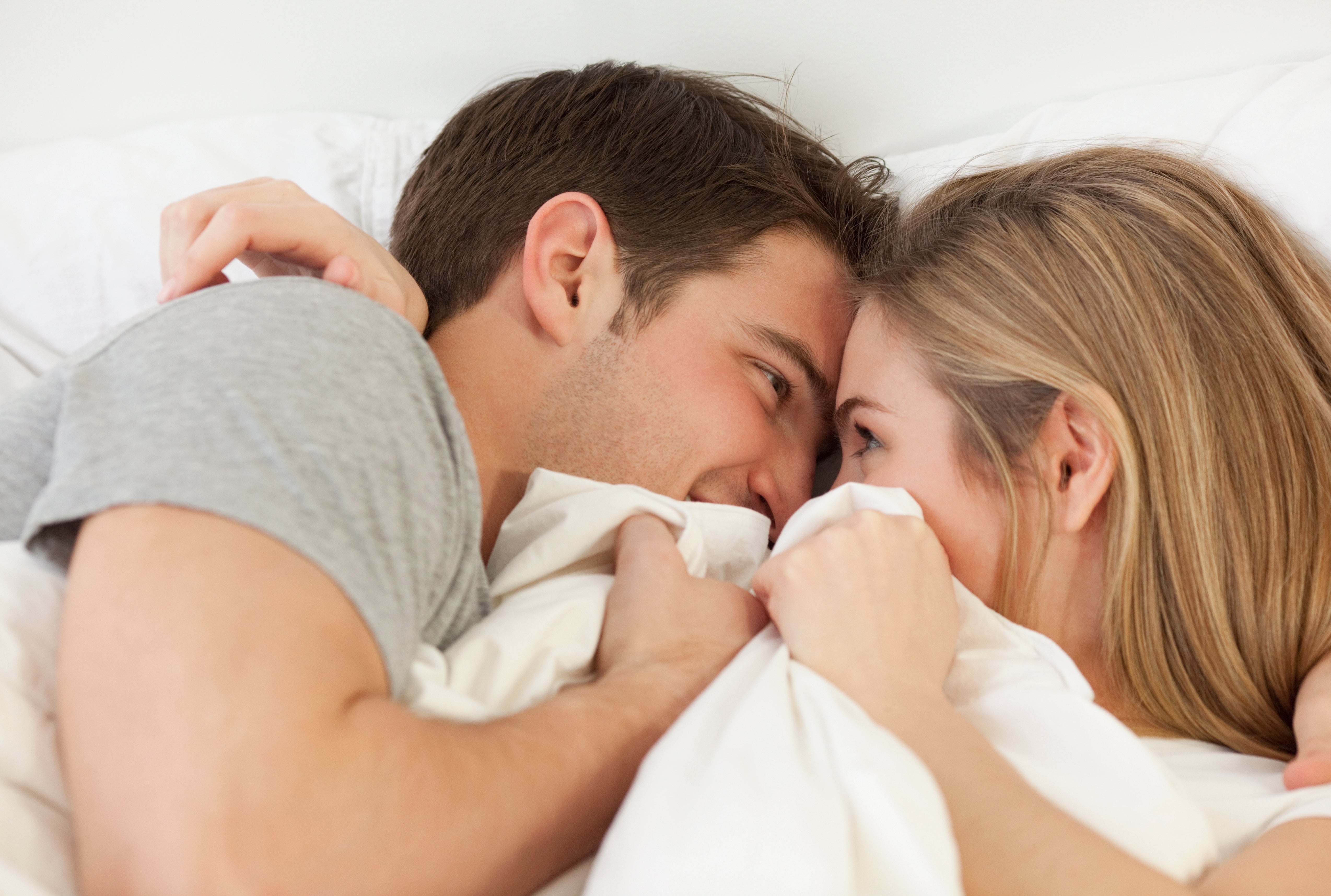 A couple snuggles under a white duvet, smiling and looking into each other’s eyes, creating a cozy and intimate atmosphere
