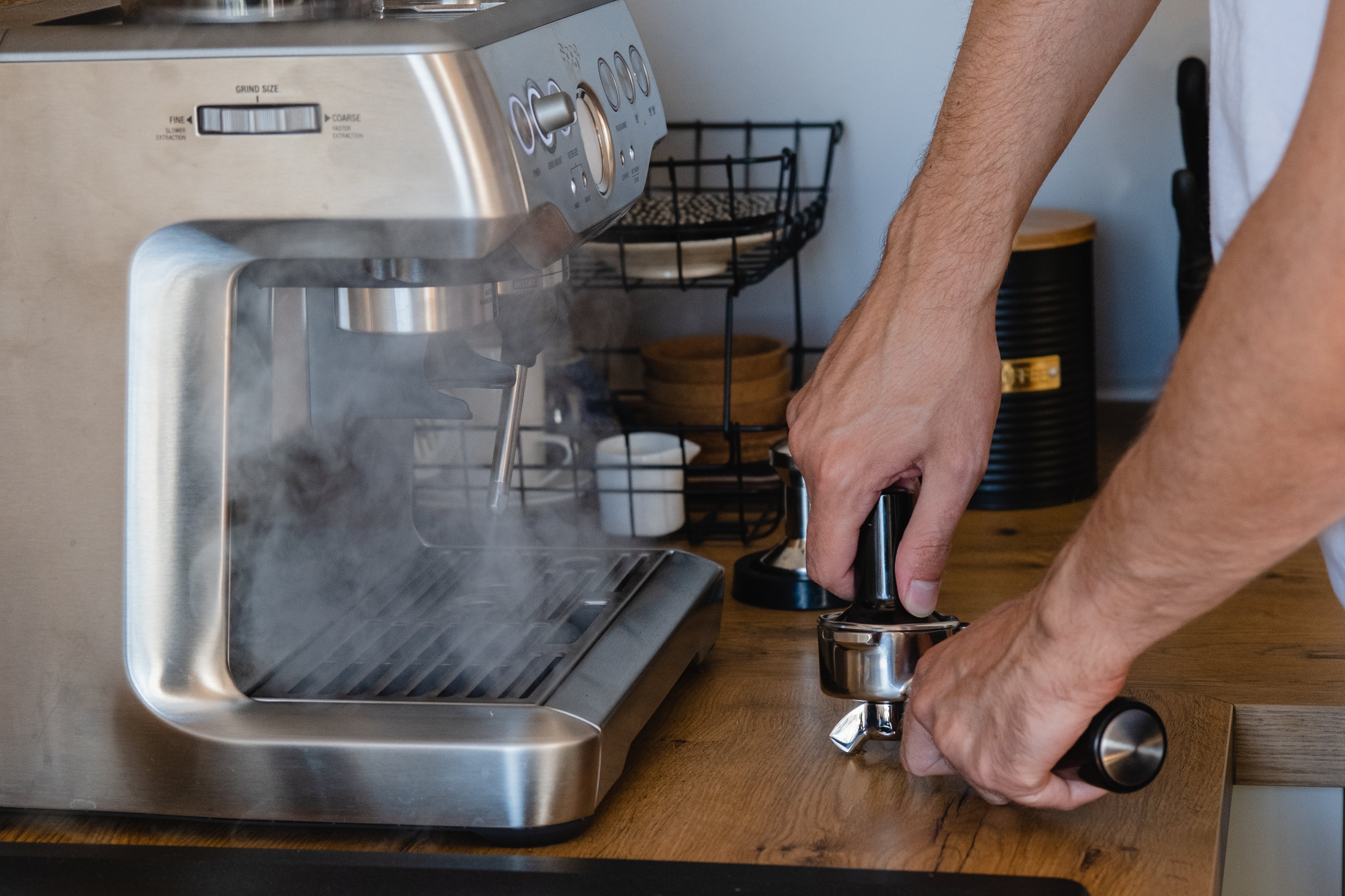 A person prepares espresso by tamping coffee grounds next to a steaming espresso machine on a wooden counter