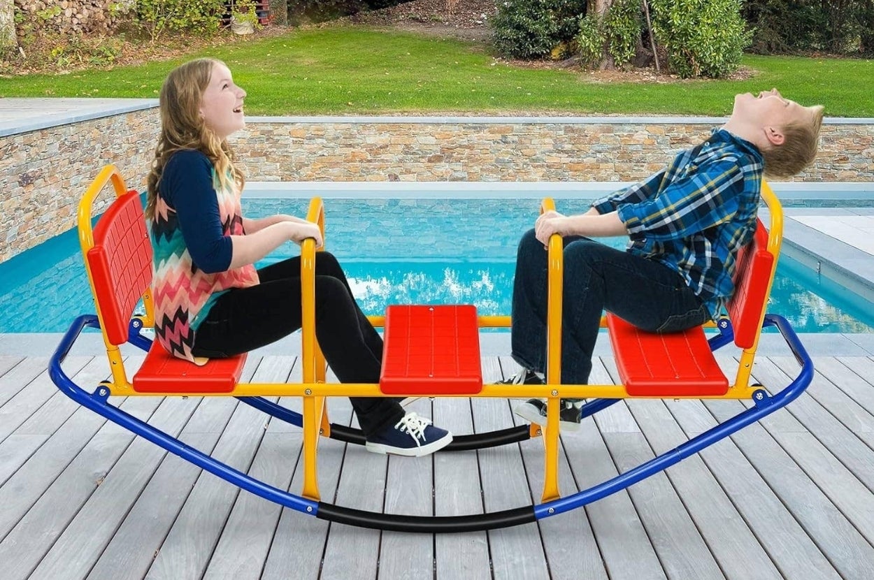 Two people in casual clothes sit on a colorful seesaw by a pool, enjoying a playful moment