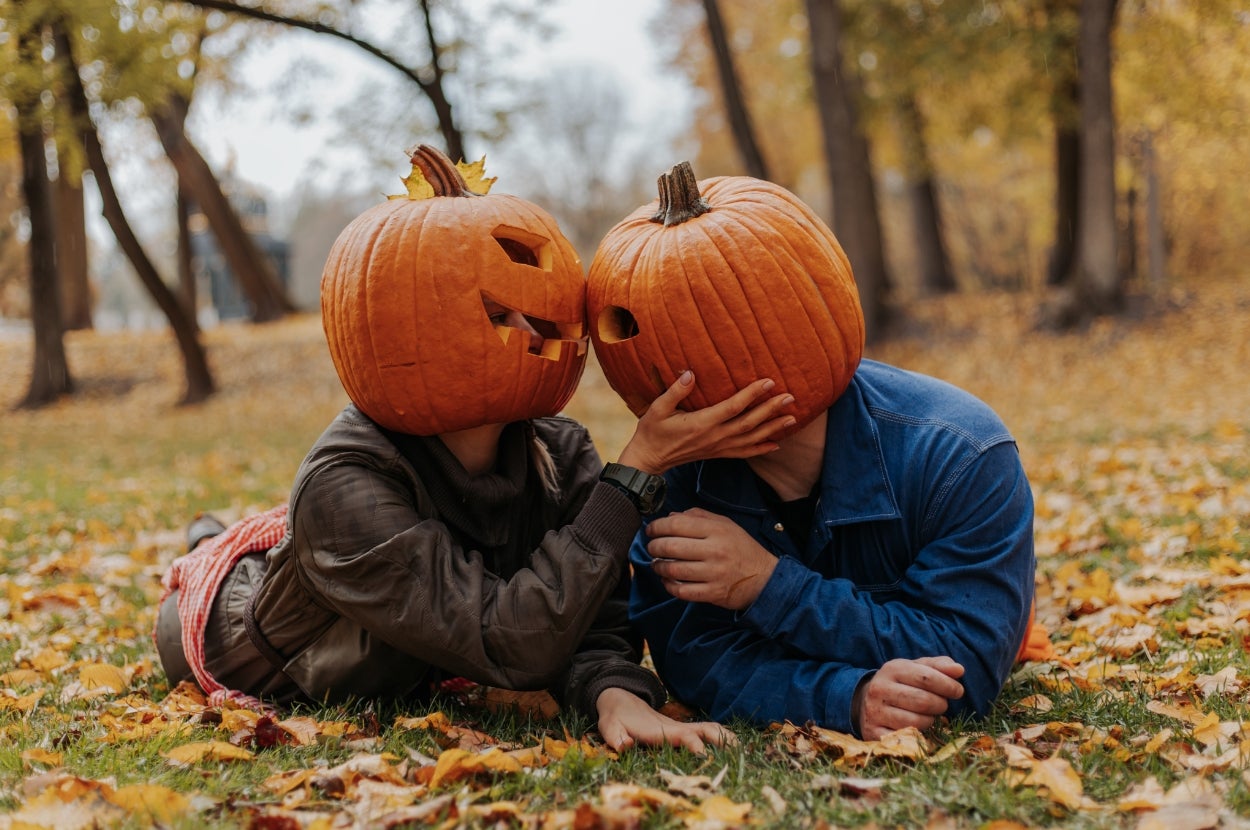 Two people lying on grass, each with a carved pumpkin over their head, as though kissing. Autumn leaves are scattered around
